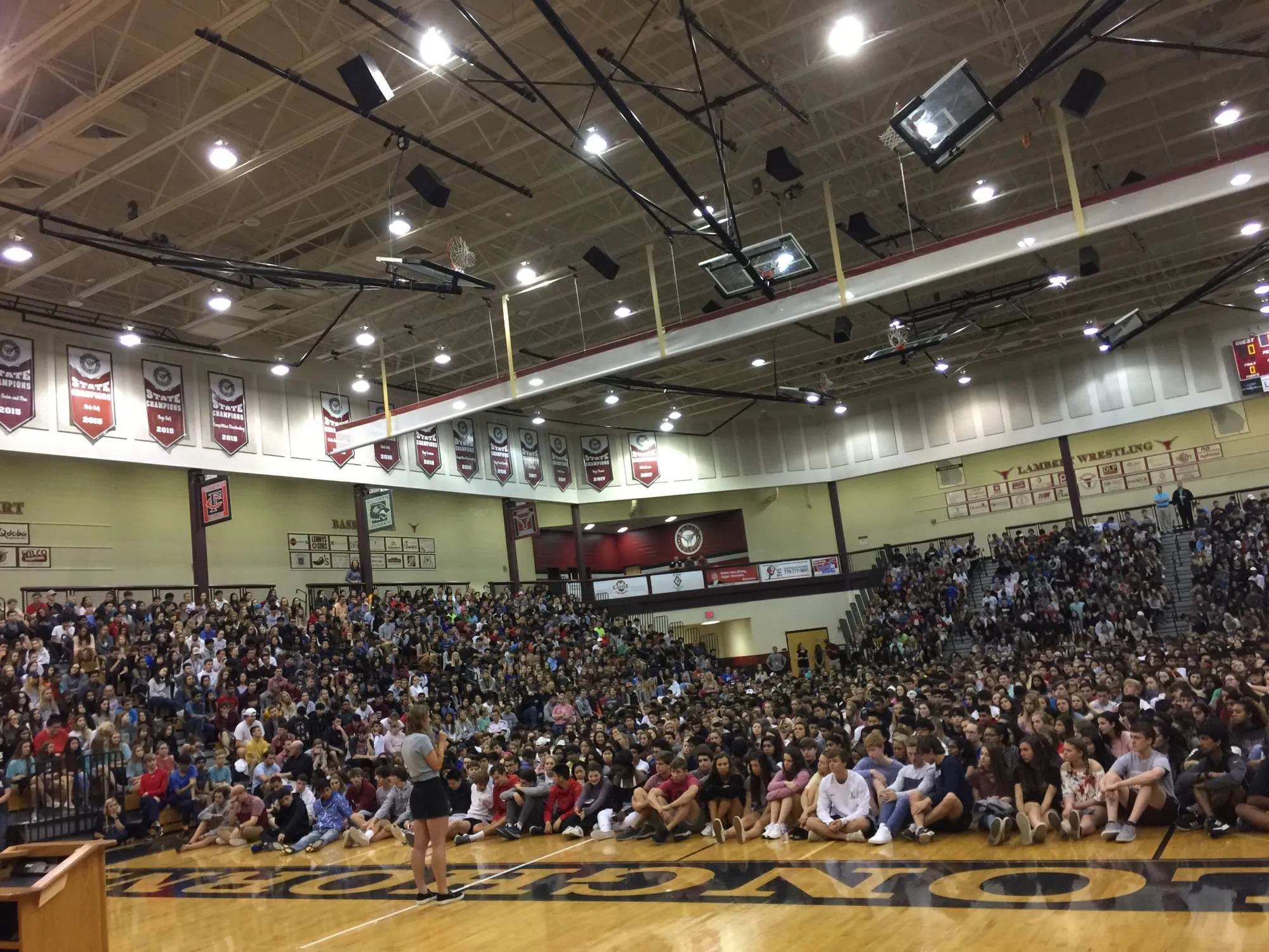 A Whisper speaker addressing a packed school gym, students filling the bleachers and sitting across the floor